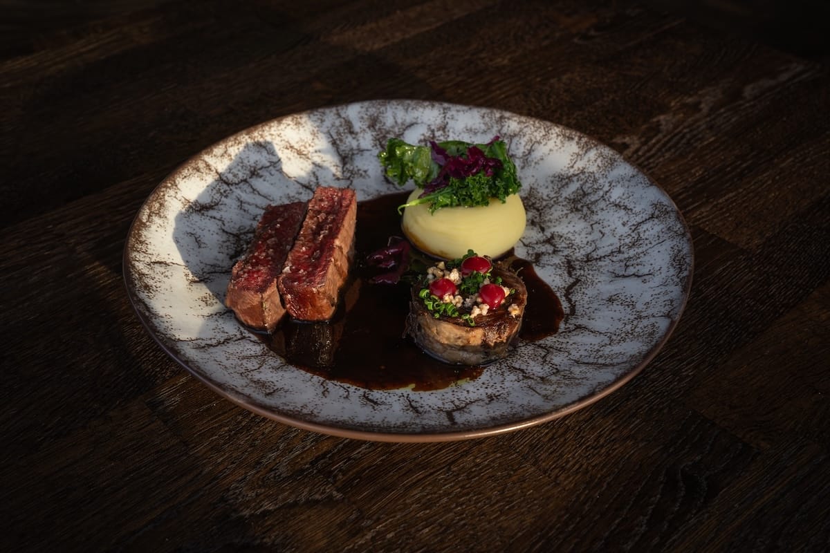 A plate with a reindeer sirloin, a reindeer entrecote wrapped with lingonberry on top, mashed potatoes topped with kale, and a game sauce.