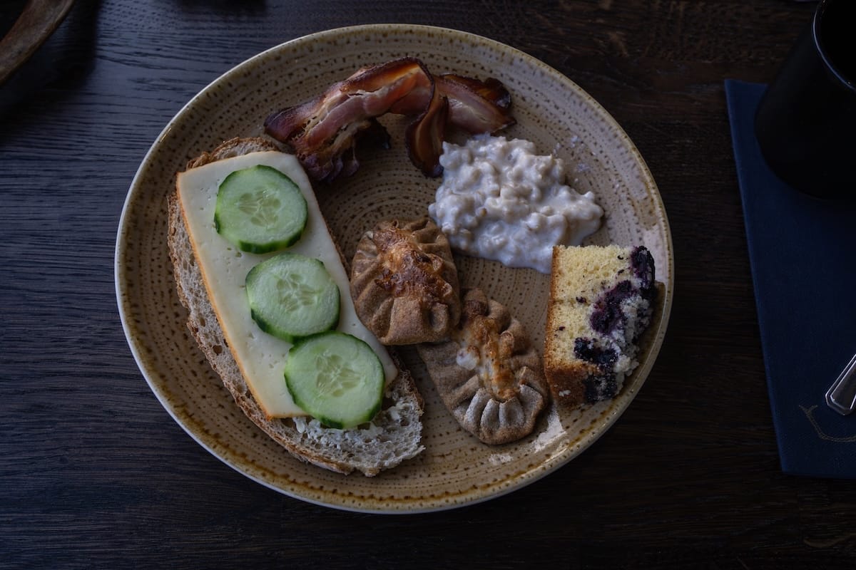 A plate of food from breakfast: bread topped with cheese and cucumbers, a karelia pie, a blueberry cake, porridge, and bacon